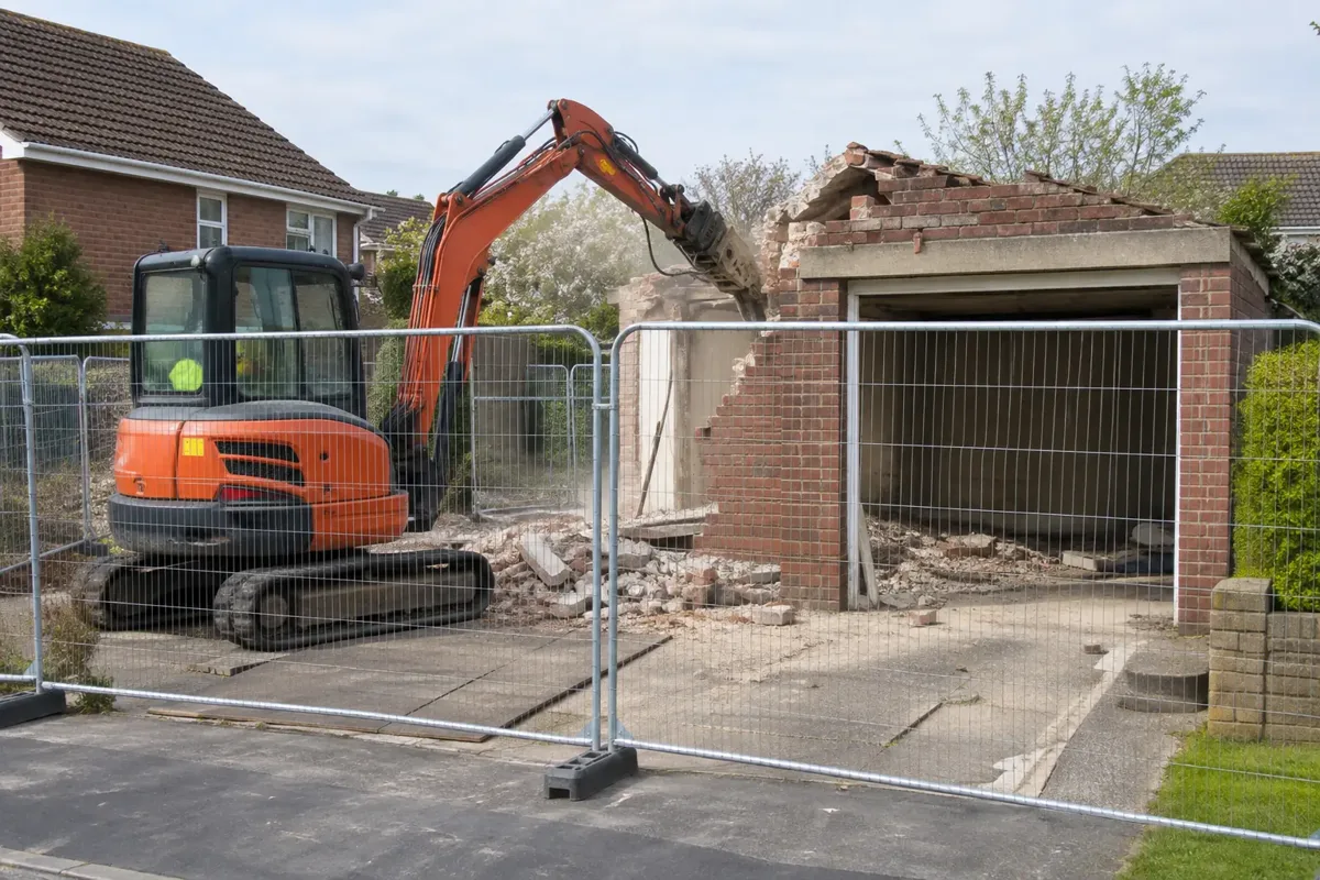 Small excavator carefully dismantling the remaining brick wall of an empty detached domestic garage, viewed outside a solid safety barrier. The garage door opening and concrete base remain recognisable, tidy cordoned UK driveway, light dust suppression, no people near machinery.